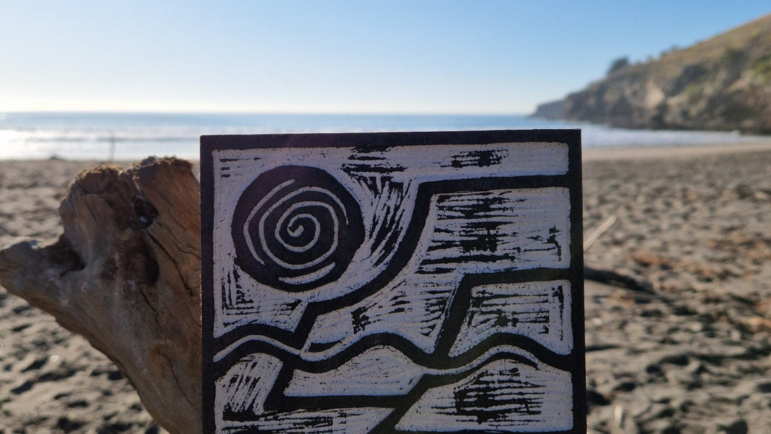 A Lino Cut block rests on a piece of driftwood on a beach
