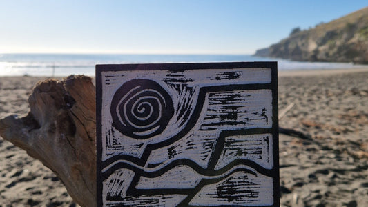 A Lino Cut block rests on a piece of driftwood on a beach