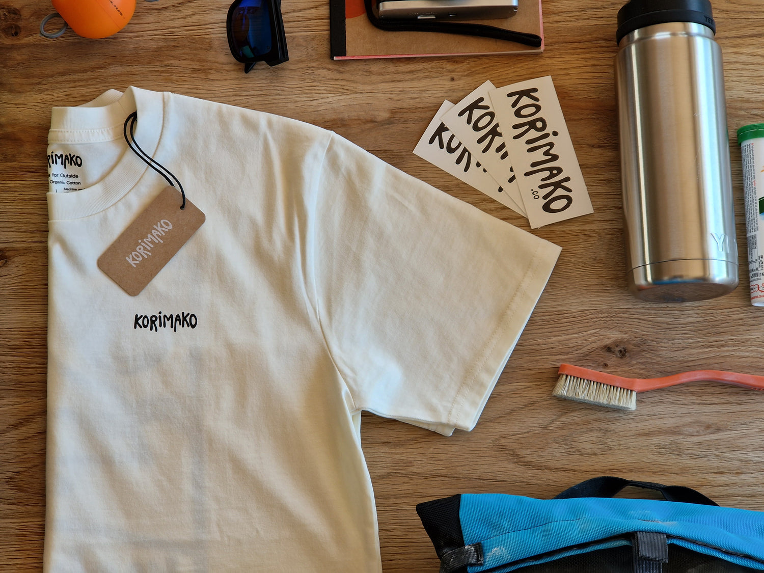 A cream-colored short-sleeve t-shirt with 'korimako' printed in black on the chest, laid flat on a wooden table surrounded by various lifestyle items like a stainless steel bottle, sunglasses, and a climbing brush.