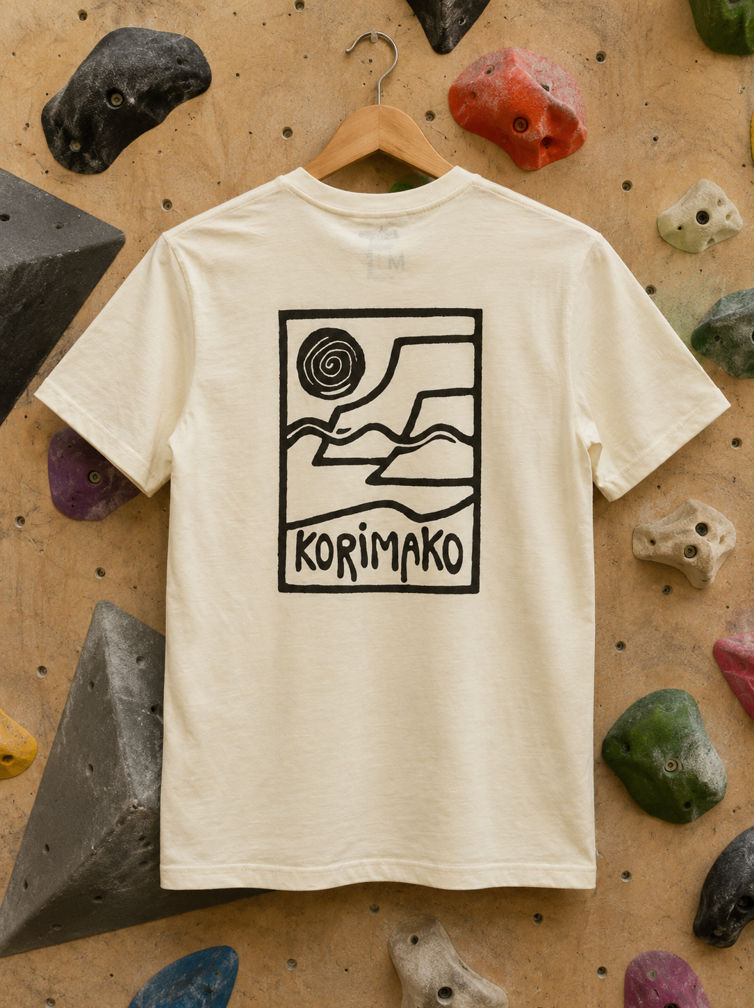 An off-white short-sleeve t-shirt with a black graphic print of a mountain landscape and the word 'KORIMAKO' hanging on a wooden hanger against a climbing wall backdrop.
